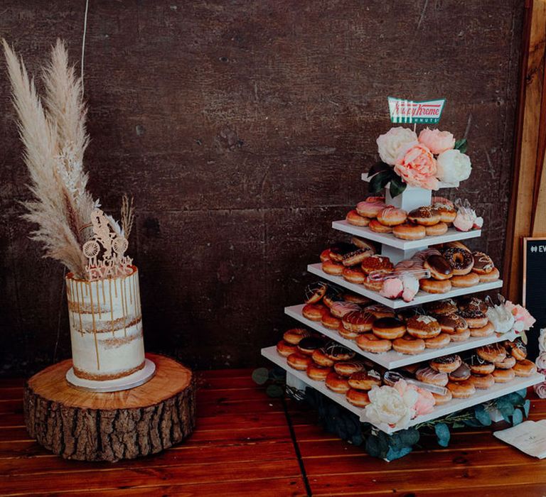 Tall semi-naked drip wedding cake on a tree slice with pampas grass and a Krispy Kreme Doughnut tower on the wedding cake table at Elmore Court 