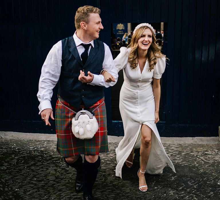 Bride in buttoned puff sleeve wedding dress with pearl headband and groom in shirt and tie with red, blue and green kilt 