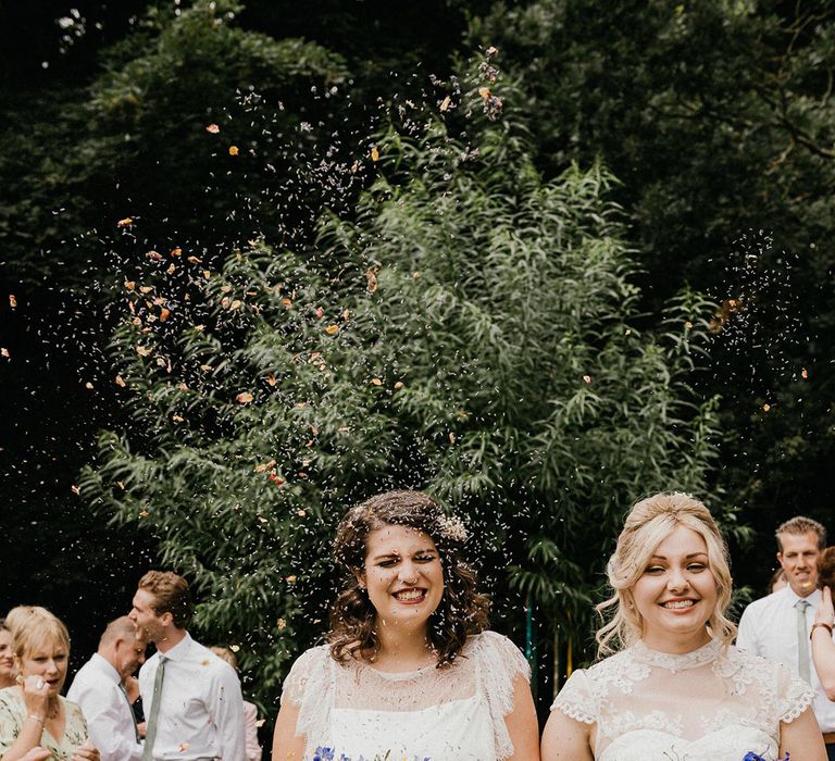 Smiling brides walk through confetti together as a married couple with sunflower, gypsophila and bellflower bouquets