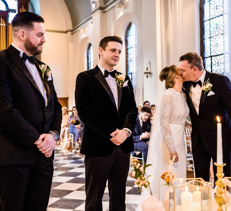 Father of the bride kisses the bride on the cheek as he walked her to the altar with groom and best man