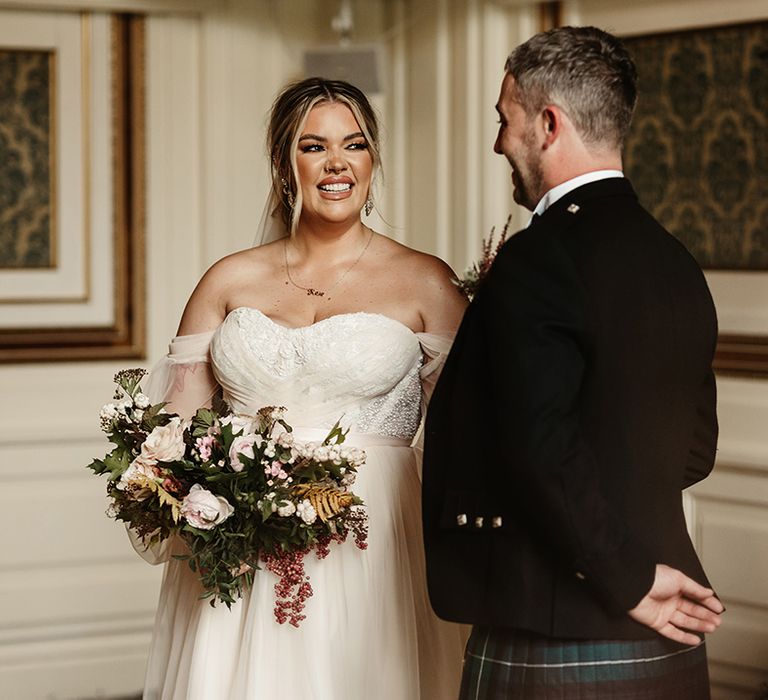 Bride and groom smile at each other with bride in custom wedding gown with sheer sleeves and autumnal classic wedding bouquet 