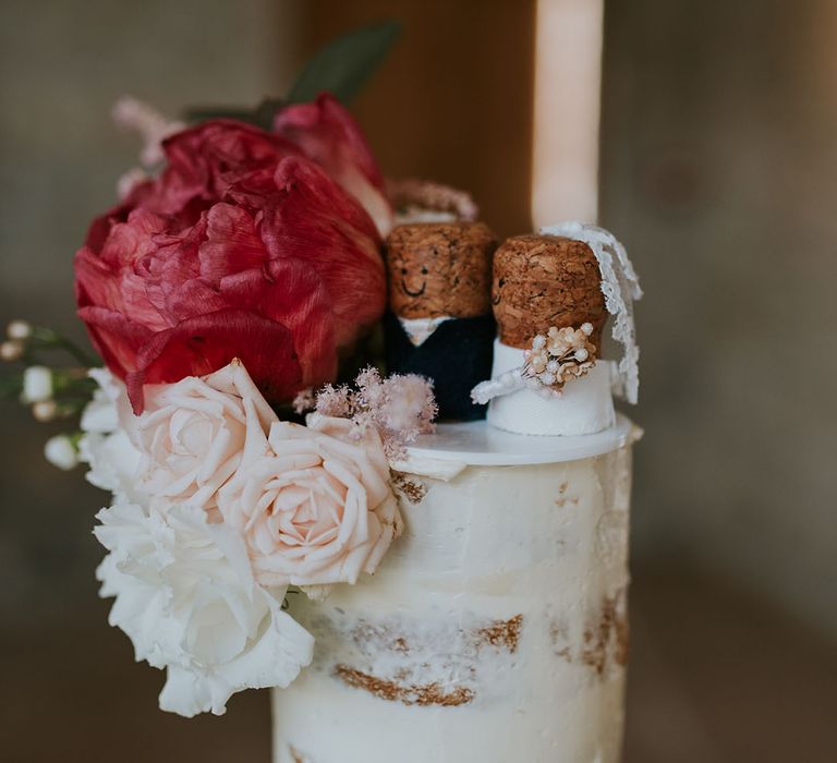 Semi-naked white frosted cake with pink and red flowers and mini cork cake toppers dressed like a bride and groom