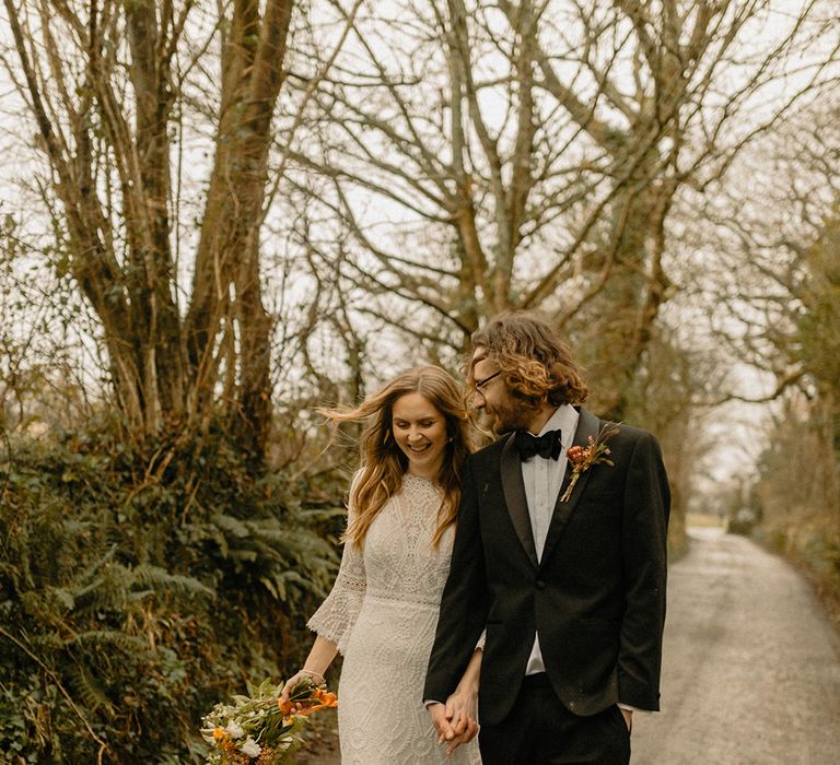 Bride and groom walk hand in hand together as they walk around the grounds of the wedding venue in black tie and lace gown