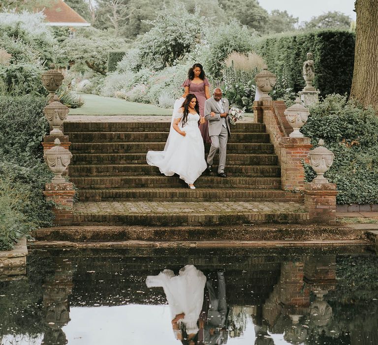 Father of the bride and bride descend the steps together as the bridesmaid in vintage style dress holds the bride's gown 