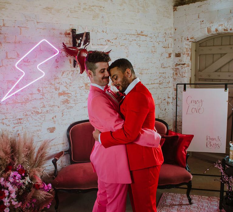 Groom in a red suit embracing his partner in a pink suit next to a pink neon lightning bolt sign and a velvet sofa 