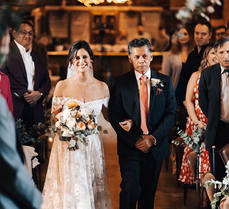 Father of the bride in black suit and coral coloured tie walks the bride down the aisle in floral lace wedding dress