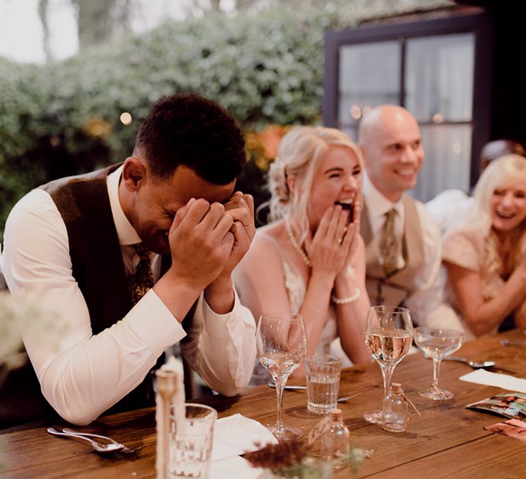 Groom covers his face with his hands as bride and other wedding guests laugh