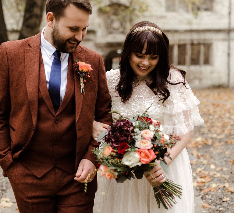 Bride and groom look down together as they smile with bride wearing sequinned Needle and Thread bridal separates and groom in brown suit