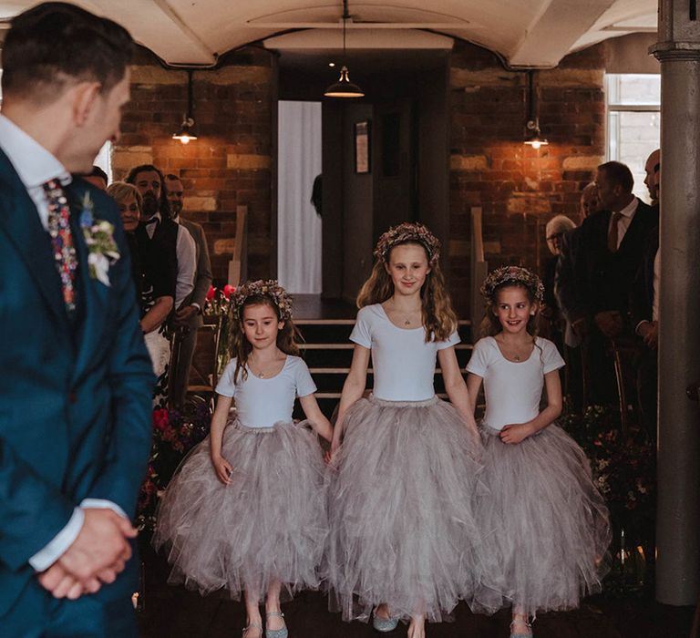 Three flower girls wearing white t-shirts and grey tulle ruffle skirts walk down the aisle together wearing matching flower crowns