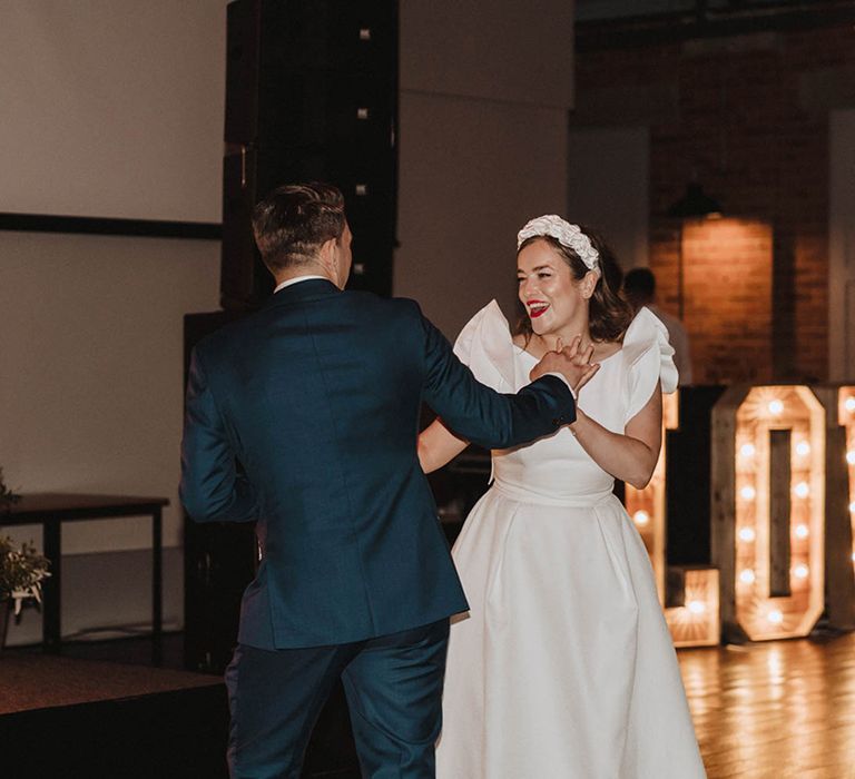 Groom in blue suit has first dance with bride in Jesus Peiro wedding dress and white ruffle headband with light up sign and colourful flowers 