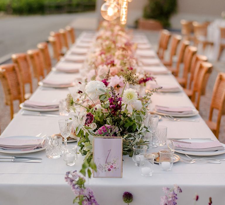 Head table complete with Edison bulbs installation and purple floral bouquets and lavender napkins on white plates and white tablecloth 