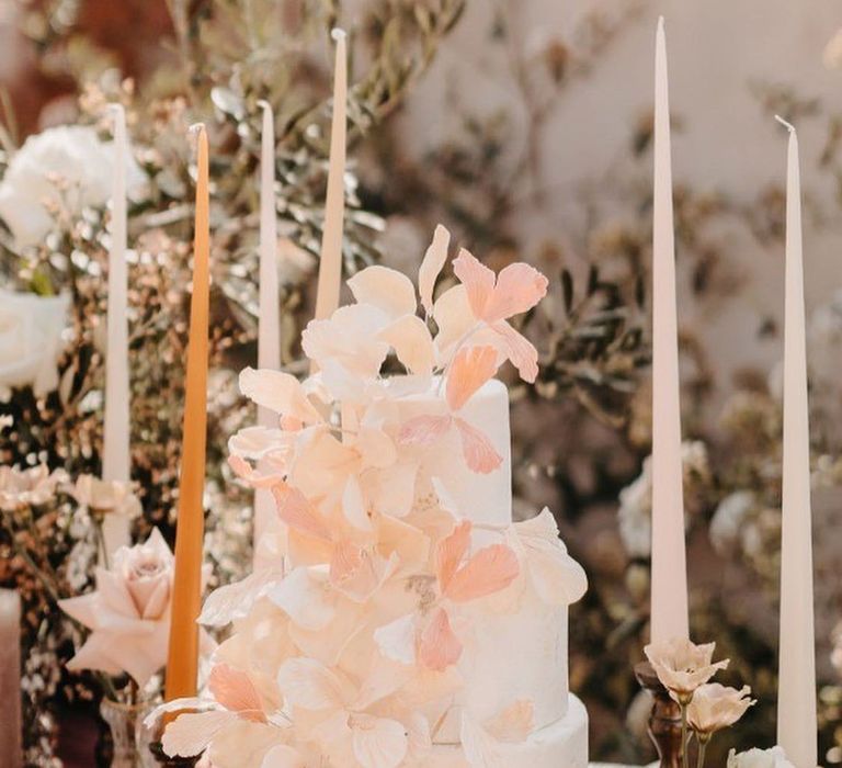 Three tier white wedding cake with peach and pink flowers and tall taper candles 