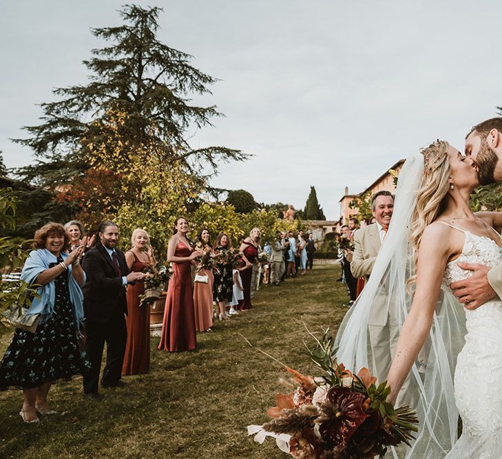 Groom kisses his bride whose Made With Love Bridal blows in the wind as wedding guests watch on after wedding ceremony outdoors