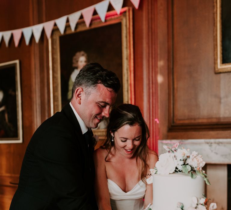 Bride & groom cut three tier wedding cake with white icing and decorative florals 