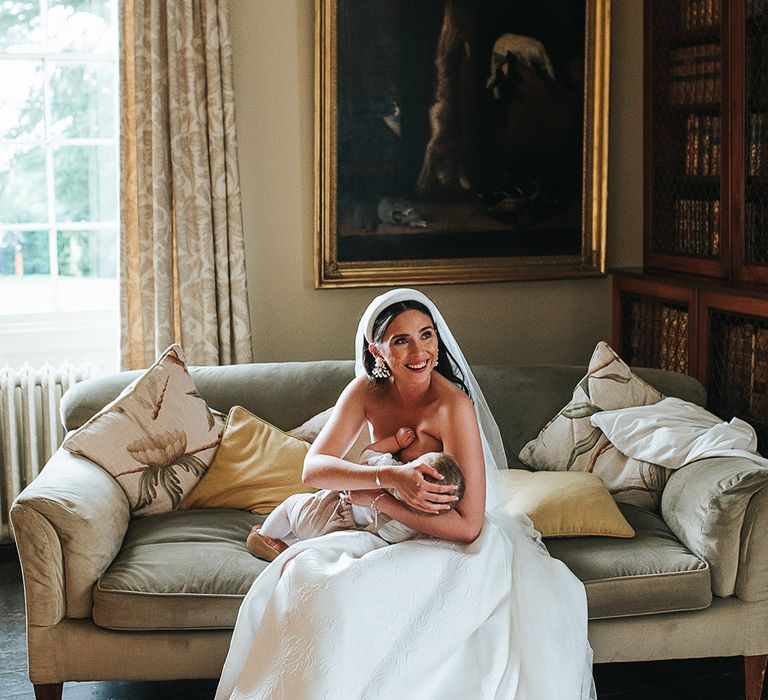 Bride wearing a chunky white headband and strapless wedding dress breastfeeds her baby boy 