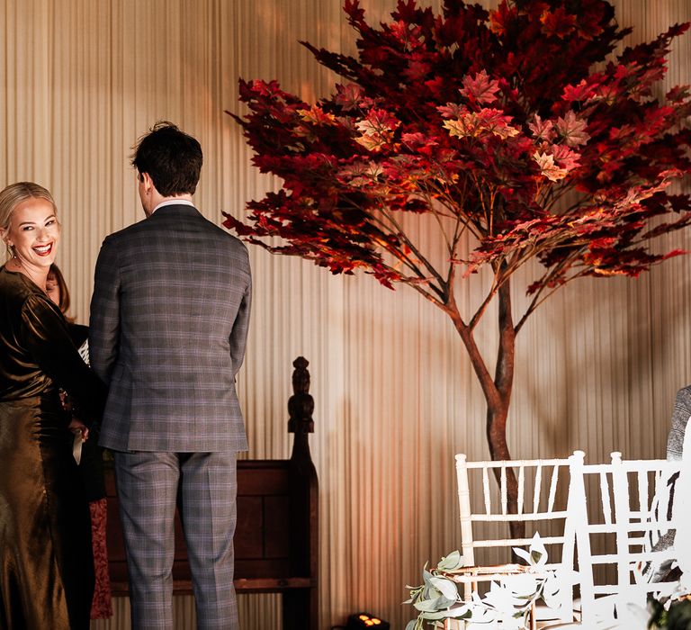 Bride wearing red lipstick looks behind her during wedding ceremony stood beside her groom and Autumnal styled tree 