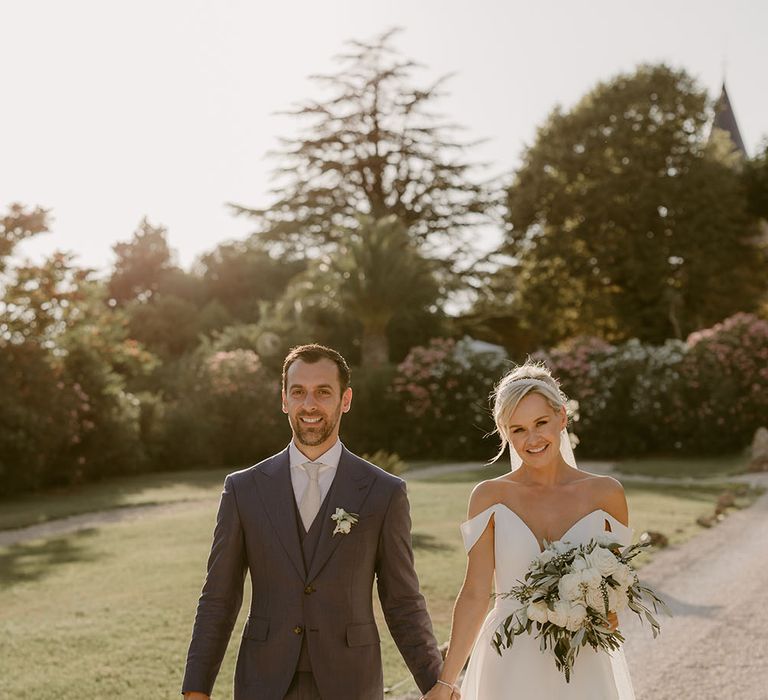 Groom wears three piece suit and holds his brides hand as they walk through the grounds of Chateau de Robernier after wedding ceremony