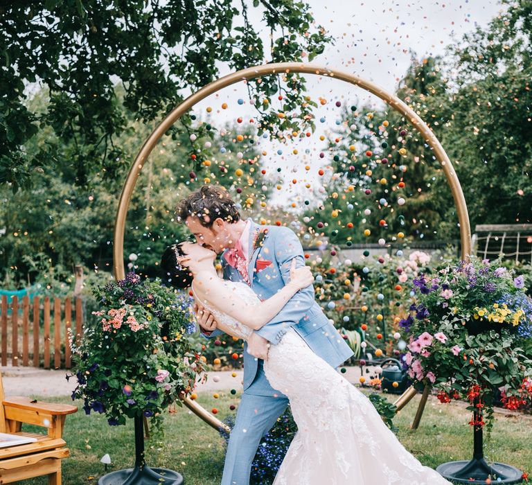 Groom dips his bride for a kiss as colourful confetti falls around them and pom pom wedding decorations hang from gold circular arch