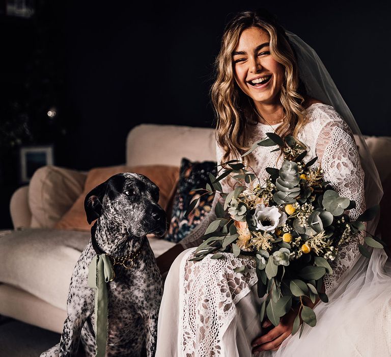 Bride laughing in a long sleeve lace wedding dress holding a yellow and white bouquet with her pet dog 