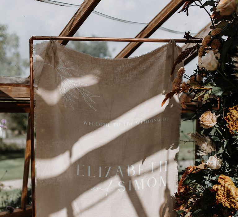 Neutral fabric welcome wedding sign on a copper frame with rust and warm toned florals 