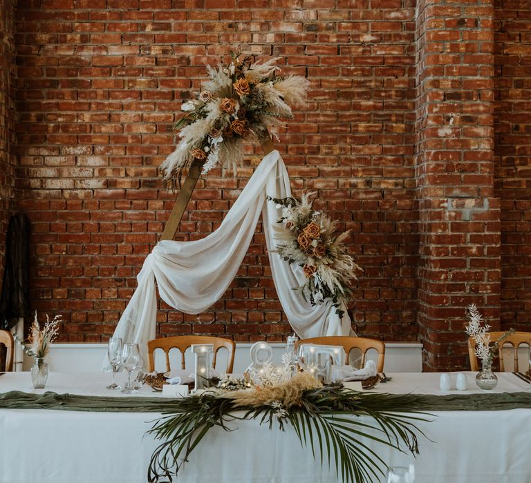 Boho rustic wedding tablescape at The Green Cornwall with large wooden triangle arch, dried flower and burnt orange and toffee rose arrangements and decor, bottle green table runner and small silver vases