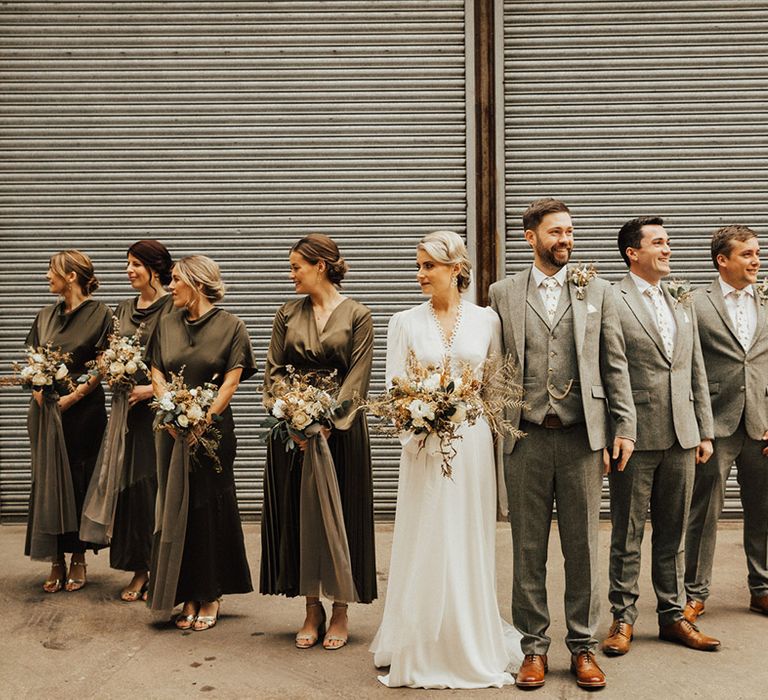 Bride & groom stand with bridesmaids in green bridesmaid dresses with long sleeves and groomsmen in grey suits 