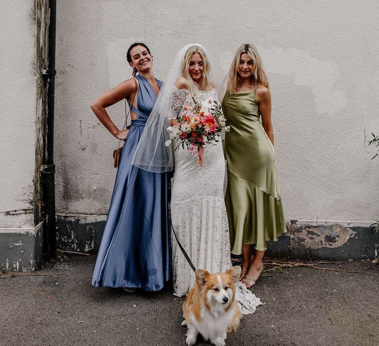 Bride in boho two piece lace bridal outfit with large colourful mixed bouquet standing with two bridesmaids in mismatched satin dresses