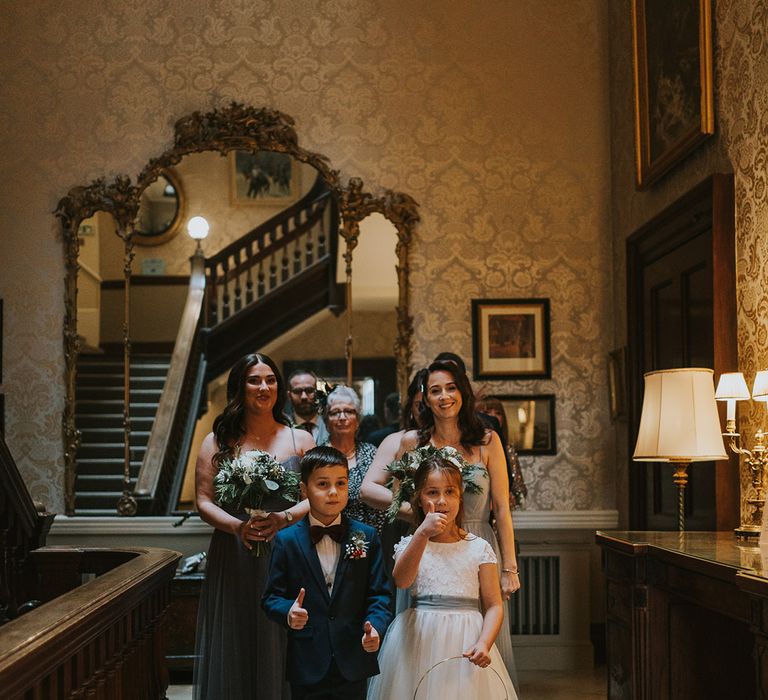 The flower girl stands with the page boy and bridesmaids in grey dresses 