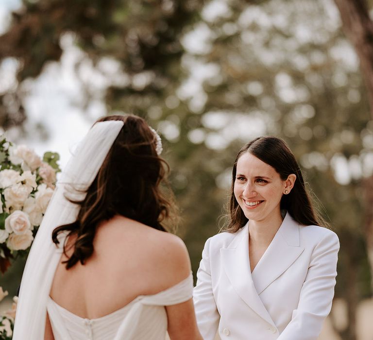 Bride in a white double breasted jacket stands at the altar holding hands with the bride for their outdoor ceremony 