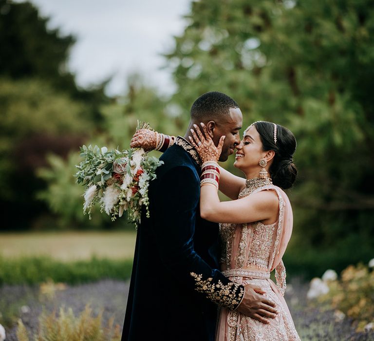 Panjabi Bride and Jamaican Groom pose at multicultural wedding. The bride wears a golden bridal Lehnga and carries a champagne bridal bouquet and the groom wears a gold embellished Indian wedding outfit