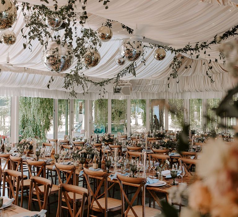 Marquee reception decorated with foliage and disco balls with simple table setting on long wooden banquet tables 