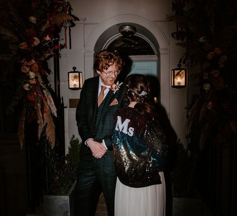 Bride in long vintage wedding dress with sparkly personalised multicoloured wedding jacket looking at groom in dark suit with orange tie at wedding party 