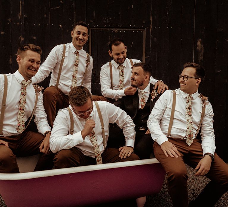 Grooms party in matching brown trousers, light brown suspenders and flower patterned ties sitting in copper bathtub at The Giraffe Shed