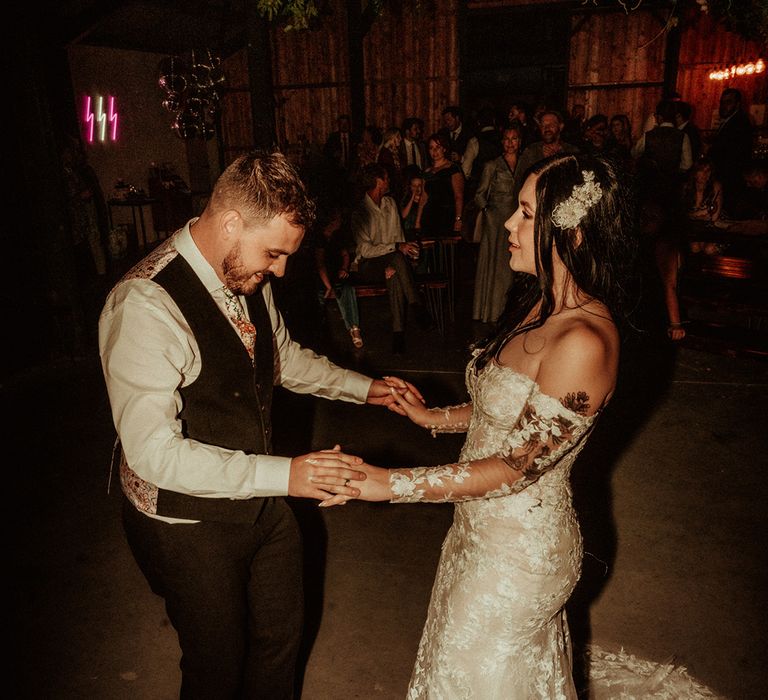 Bride in off the shoulder bodycon wedding dress with long, sheer sleeves and lace detailing holding hands and dancing with groom in black waistcoat with patterned back and flower patterned tie 