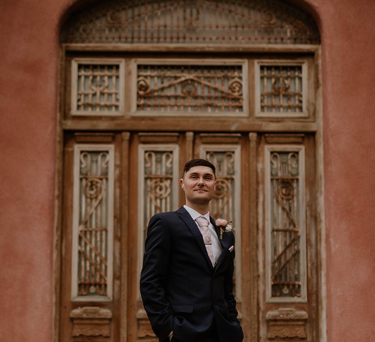 Groom wears blue three piece suit complete with pale pink tie, pocket square, brogues and floral buttonhole 