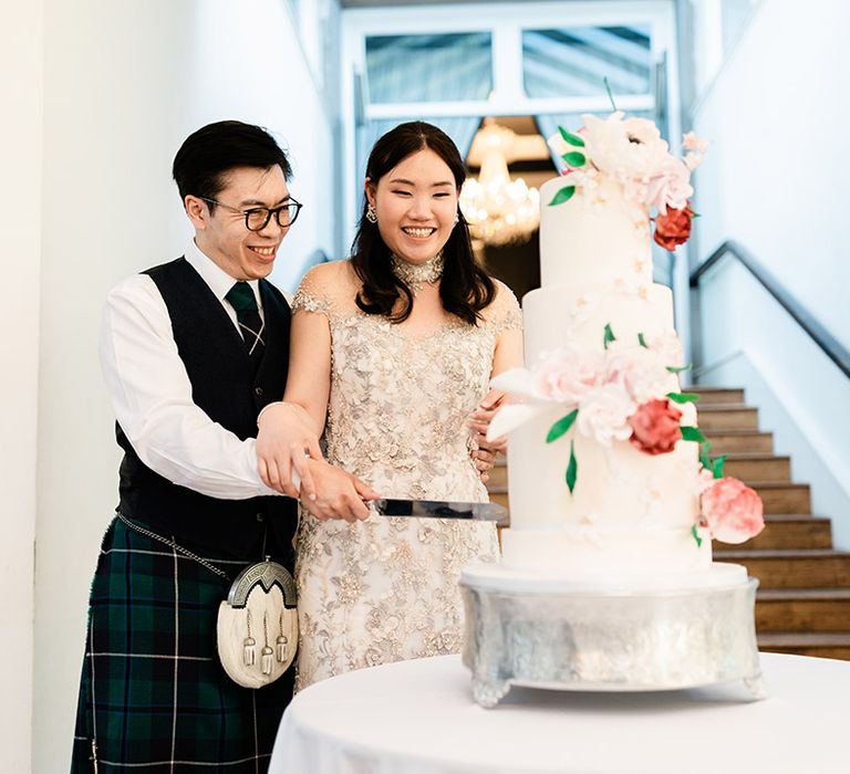 Bride in bespoke reception dress cuts three tier wedding cake with white frosting and fondant floral decor