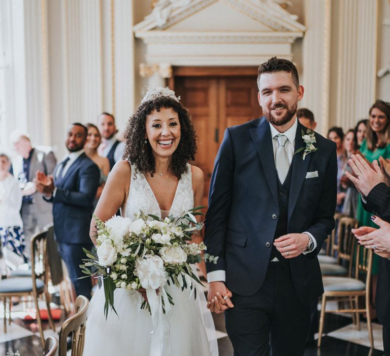 Bride carries white floral bouquet with green foliage and walks beside her groom in three piece suit 