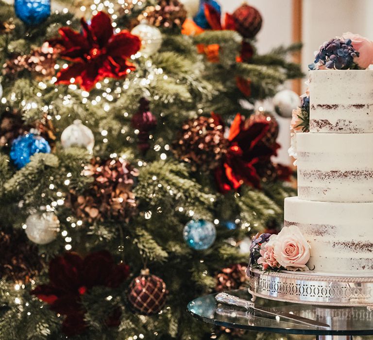 Three-tier chocolate semi-naked wedding cake with white frosting and pink rose decor in front of the Christmas tree 