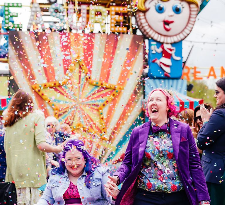 Bride in wheelchair wearing personalised denim jacket, pink wedding dress and blue tights holding hands with bride in purple velvet blazer and black suit trousers with purple bowtie, pink patterned pocket square and colourful patterned shirt walking through their colourful confetti throw 