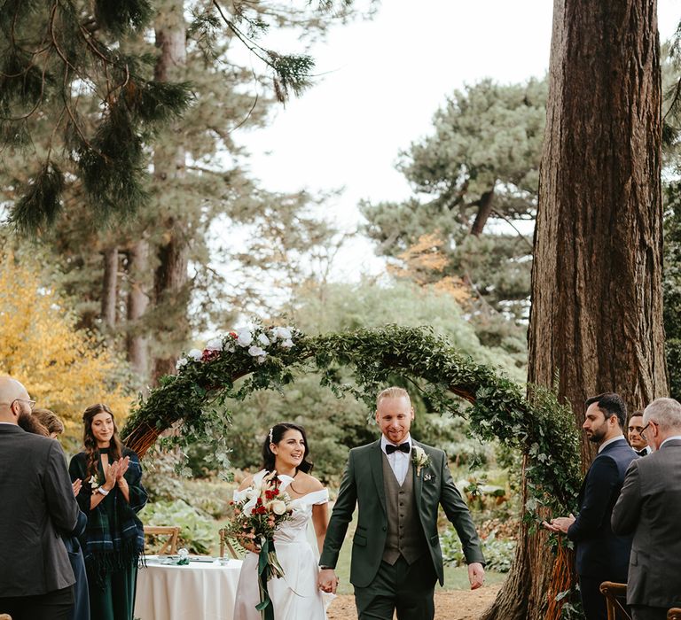 Bride carries white orchid bridal bouquet and walks alongside her groom in green three-piece suit and floral buttonhole 