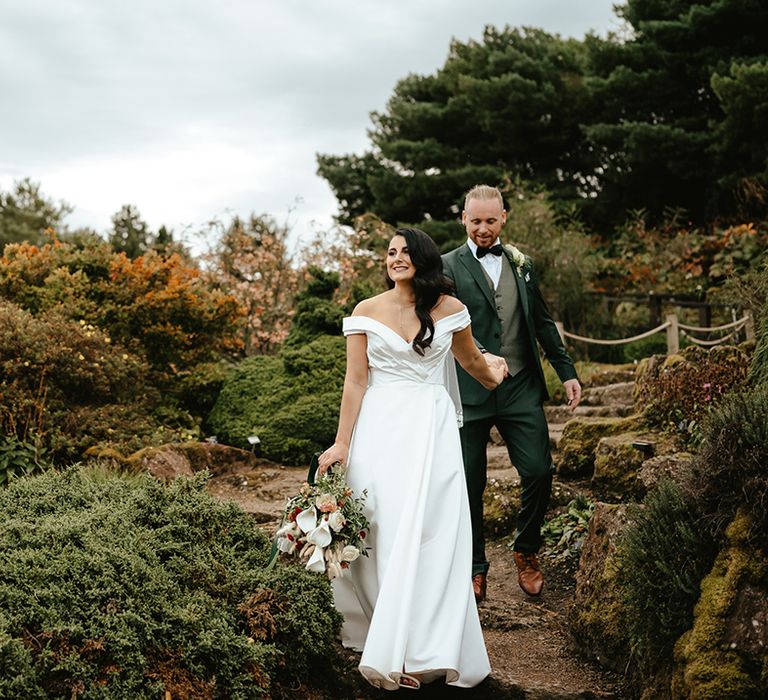 Bride in off-the-shoulder wedding dress leads her groom in green suit in botanical setting outdoors 