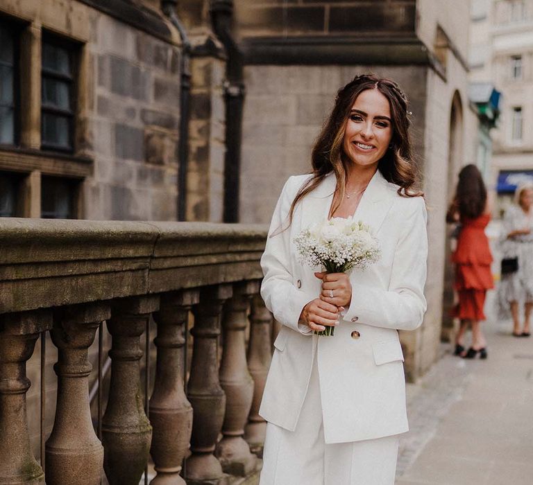 Bride wears white suit jacket complete with wide leg trousers and white rose bouquet