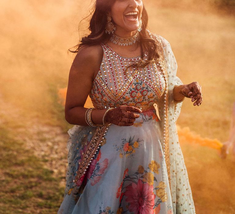 Bride in colourful lenhenga laughs and stands amongst orange smoke bomb during outdoors portraits