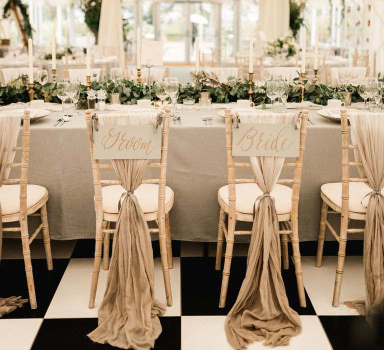 Glam white and gold marquee wedding with the wedding chairs decorated with grey sashes tied with ribbon and signage for the bride and groom's chairs 