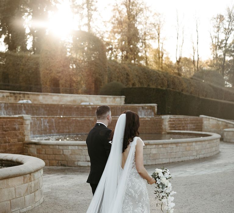 Bride wearing a veil with bow strap with watteau train in a floral embellished dress walking with the groom in the gardens at their wedding venue 