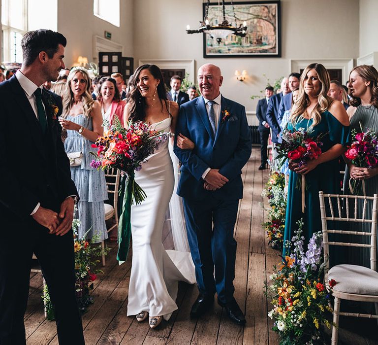 Father of the bride in a blue suit walks the bride to the end of the aisle where the groom waits 