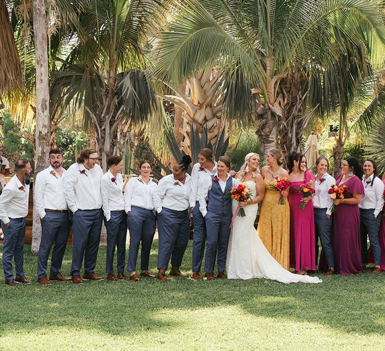 Brides stand with the colourful I do crew who wear mismatched bridesmaid dress in vibrant colours and white shirts with blue trousers 