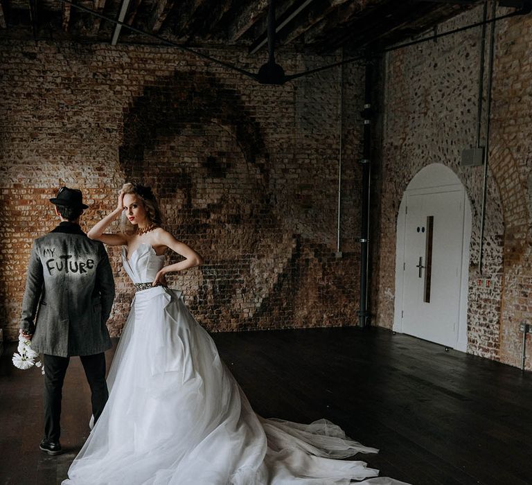 Bride in romantic organza and tulle a-line wedding day bridal ballgown with a flowing skirt, sculpted bandeau top and patterned black belt with dramatic, chunky brass necklace standing with groom in personalised dark grey jacket with "my future" graffiti style lettering, black trousers and black fedora hat 