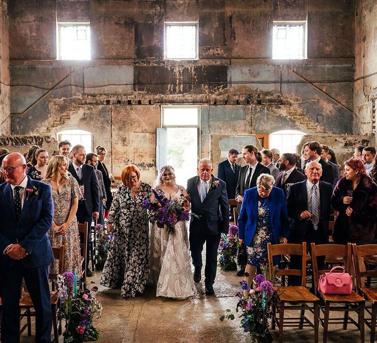 Bride in dainty leaf 3d appliqué sheer bridal gown and cathedral length veil holding bright purple bridal bouquet with purple hydrangeas, pink garden roses, purple orchids, lilac carnations, dried flowers and peacock blue feathers walking down the aisle of London Asylum Chapel with her parents  