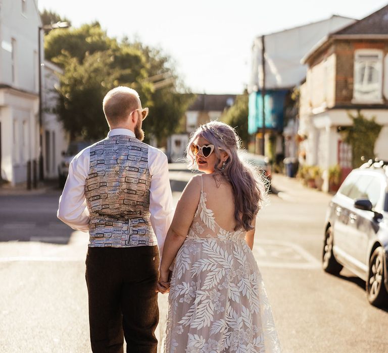 Bride in leaf appliqué sheer Evie Young bridal gown and cathedral length veil and white heart shaped sunglasses walking through London with groom in silver, brown and green waistcoat and dark trousers 